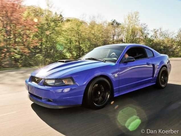 Modified blue 1999–2004 Ford Mustang New Edge coupe driving on a road, featuring black aftermarket wheels, lowered suspension, and a sporty stance.