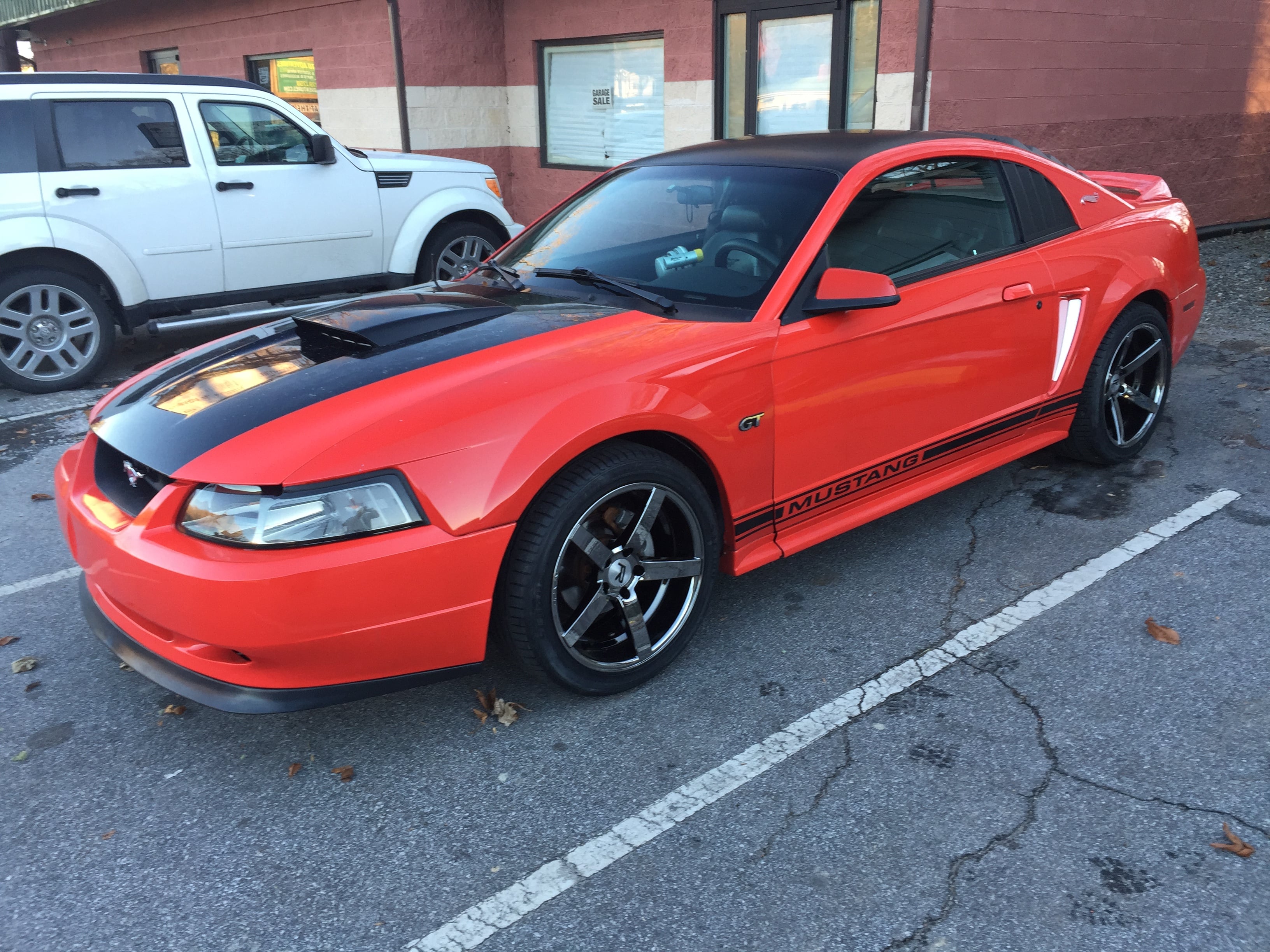 Red 1999–2004 Ford Mustang GT New Edge coupe with black hood stripe, hood scoop, and black aftermarket wheels parked in a lot.